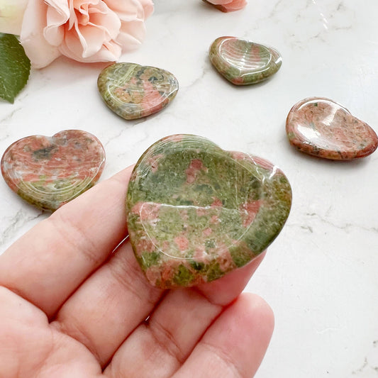 A hand holds a polished, heart-shaped Unakite Heart Shaped Worry Stone, which is green and pink in color. In the background, additional unakite hearts are scattered on a marble surface, alongside blush-colored flowers and green leaves. This scene evokes a sense of emotional balance and spiritual grounding.