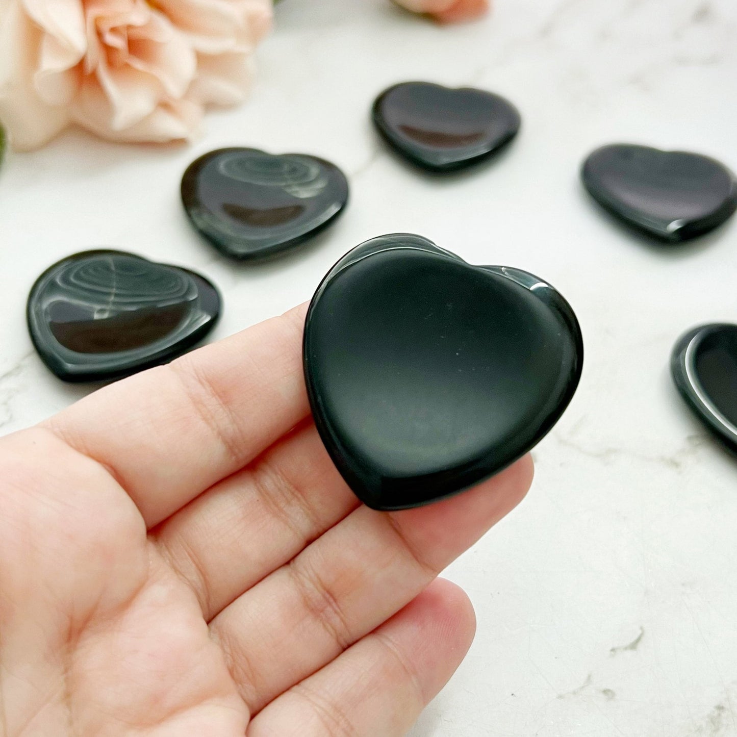 A hand holds an Obsidian Heart Shaped Worry Stone against a marble background, with several similar stones scattered around. Pale pink flowers are partially visible in the background, hinting at an atmosphere of emotional healing and stress relief.