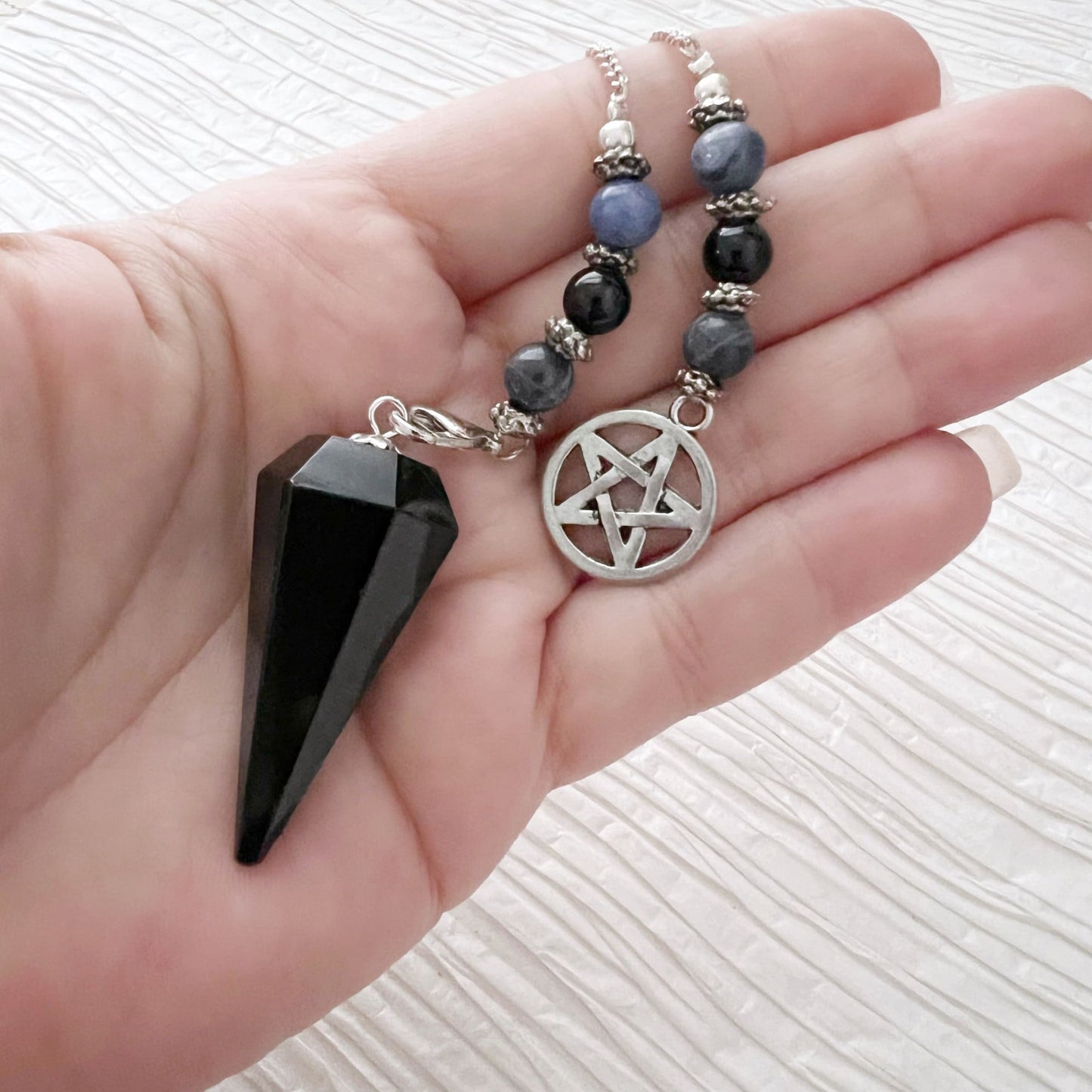 A Black Obsidian Hexagonal Pendulum with Sodalite & Pentacle Charm and a beaded chain with a silver pentagram pendant are placed on top of a closed black book. The book has gold-embossed text on the cover, partially visible, grounding energies and intuitive powers. The background features a textured white surface.