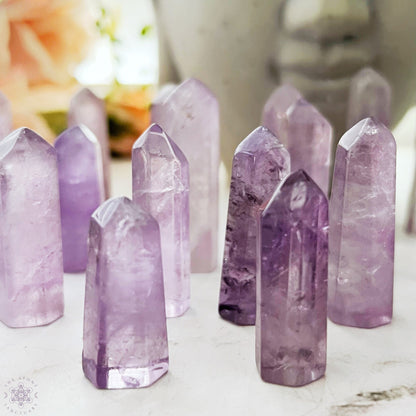 A 1-2" Polished Amethyst Point arranged upright on a smooth surface. The crystals, perfect for energy generators or crystal grids, vary in size and shape, with multiple facets catching the light and revealing the natural inclusions within the stones. A blurred background adds depth.