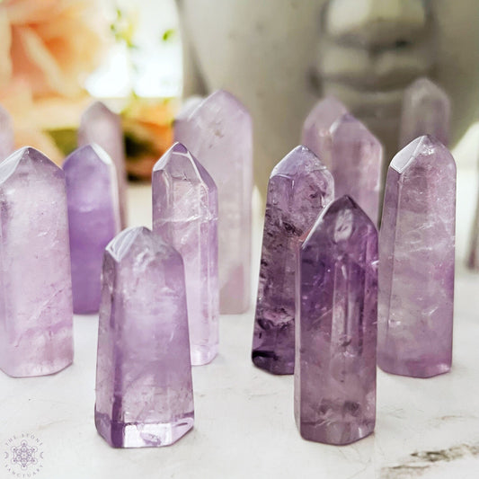 A 1-2" Polished Amethyst Point arranged upright on a smooth surface. The crystals, perfect for energy generators or crystal grids, vary in size and shape, with multiple facets catching the light and revealing the natural inclusions within the stones. A blurred background adds depth.