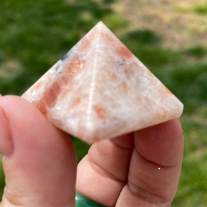 A hand with a manicured nail holds a small, polished 1" Mini Sunstone Pyramid with a mix of pink, white, and black colors. Known for bringing good fortune and self-empowerment, it is displayed against a grassy area with patches of dirt in the background.