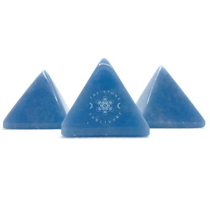 A hand holding a smooth, light blue 1" Mini Angelite Pyramid against a blurred green background. The pyramid has a shiny surface and distinct triangular faces, showcasing its polished and symmetrical structure, ideal for psychic healing.