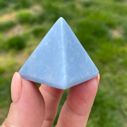 A hand holding a smooth, light blue 1" Mini Angelite Pyramid against a blurred green background. The pyramid has a shiny surface and distinct triangular faces, showcasing its polished and symmetrical structure, ideal for psychic healing.