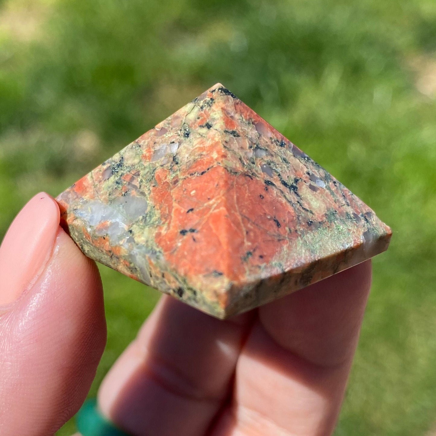 Close-up of a hand holding a 1" Mini Unakite Pyramid with a reddish-orange, green, and gray marbled pattern. The background is blurred greenery, emphasizing the stone's beauty and its potential for emotional balance.