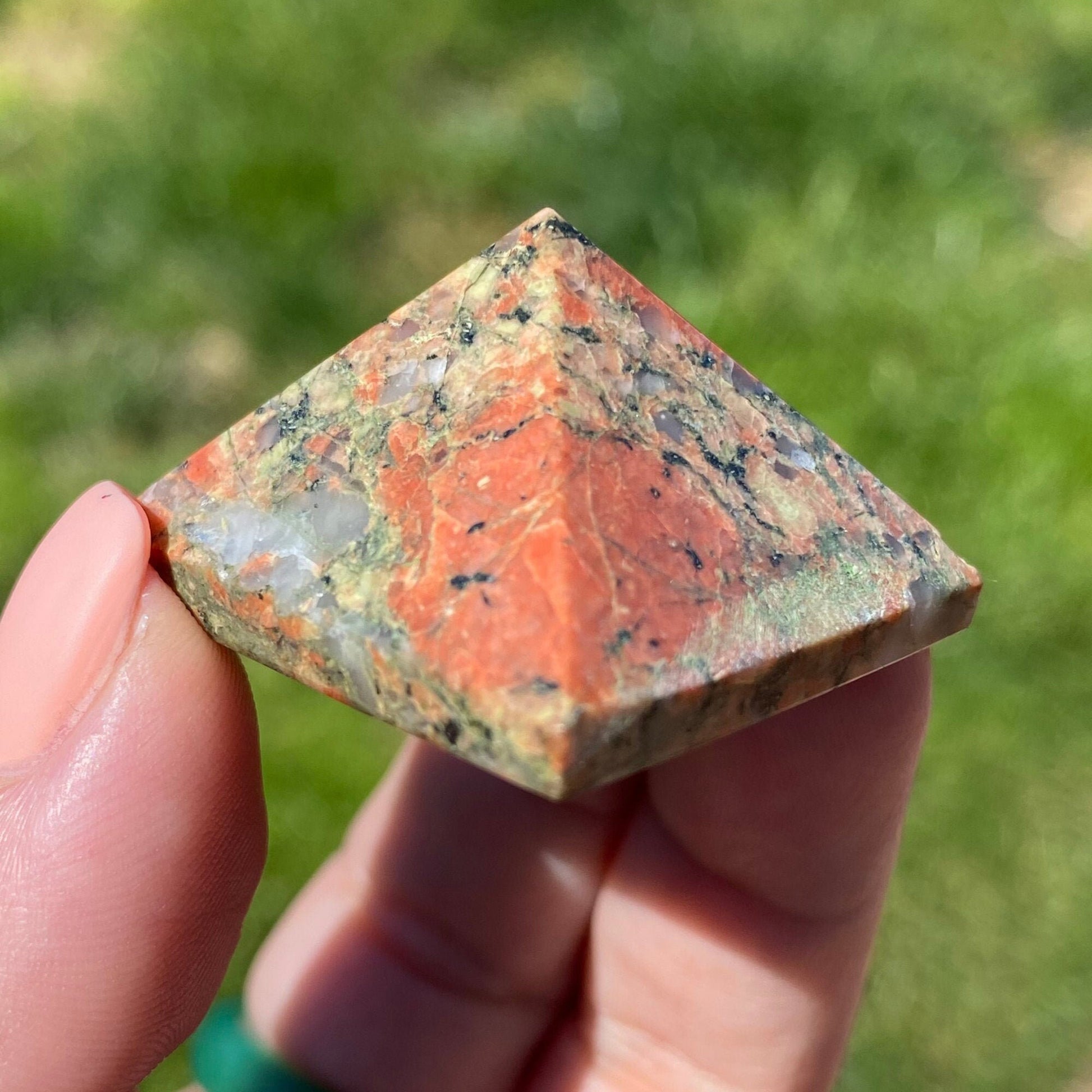 Close-up of a hand holding a 1" Mini Unakite Pyramid with a reddish-orange, green, and gray marbled pattern. The background is blurred greenery, emphasizing the stone's beauty and its potential for emotional balance.