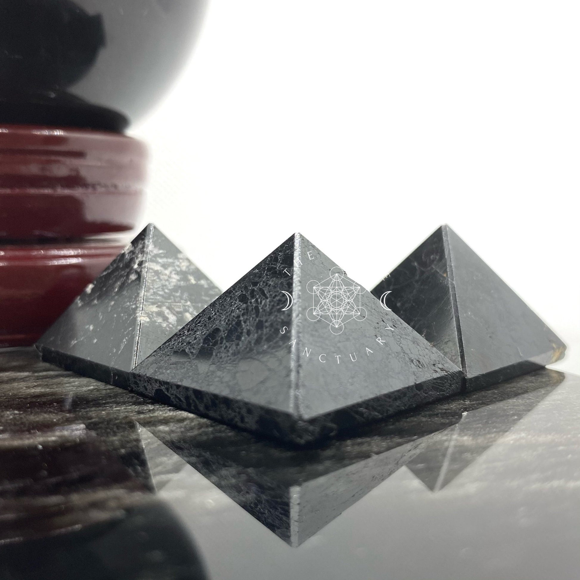 A person holds a small, 1" Mini Black Tourmaline Pyramid against a blurred background of green grass. The smooth, black stone with slight white veining is visible in daylight. Known for its protective properties, it transmits calming energy through the thumb and part of the index finger that are also visible.