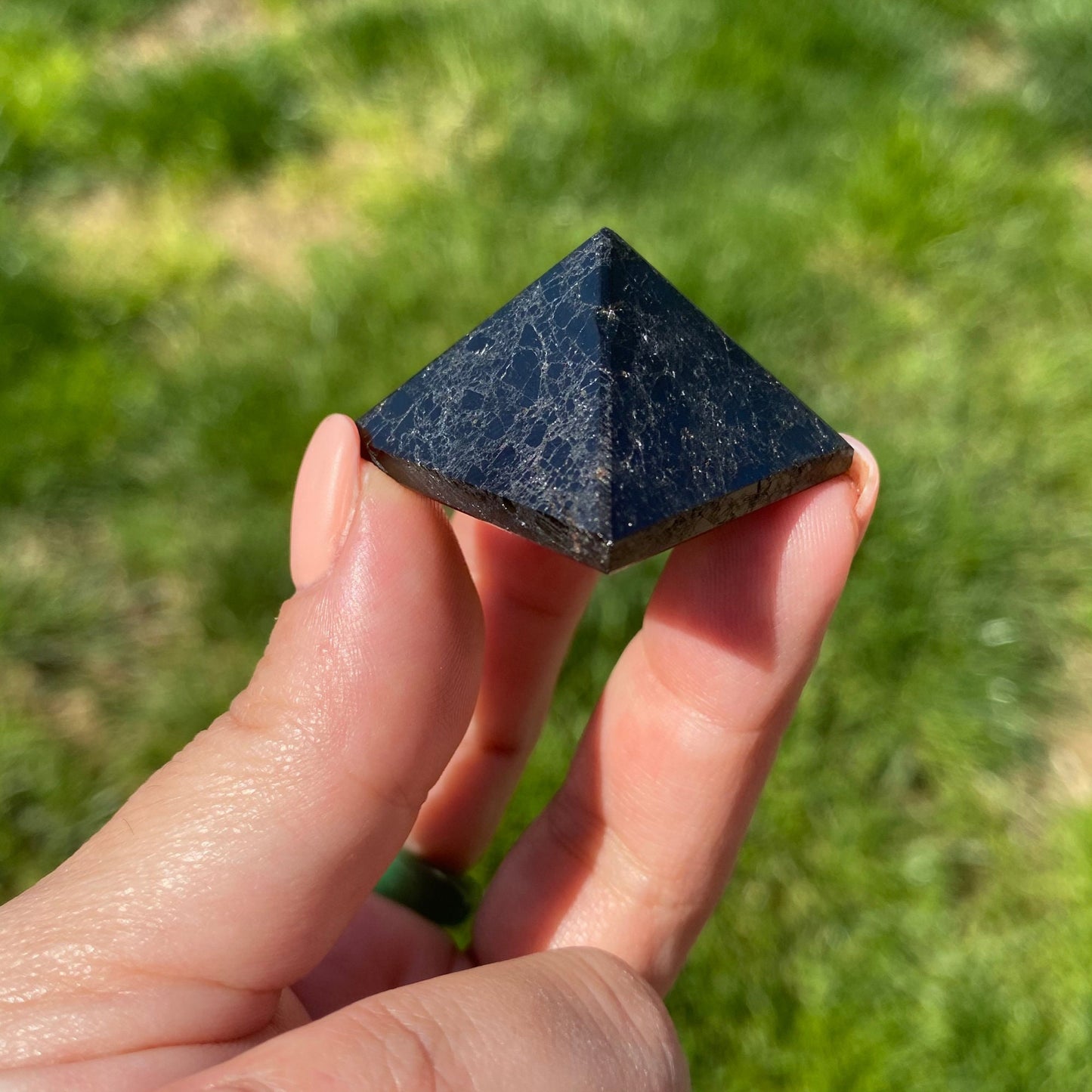 A person holds a small, 1" Mini Black Tourmaline Pyramid against a blurred background of green grass. The smooth, black stone with slight white veining is visible in daylight. Known for its protective properties, it transmits calming energy through the thumb and part of the index finger that are also visible.