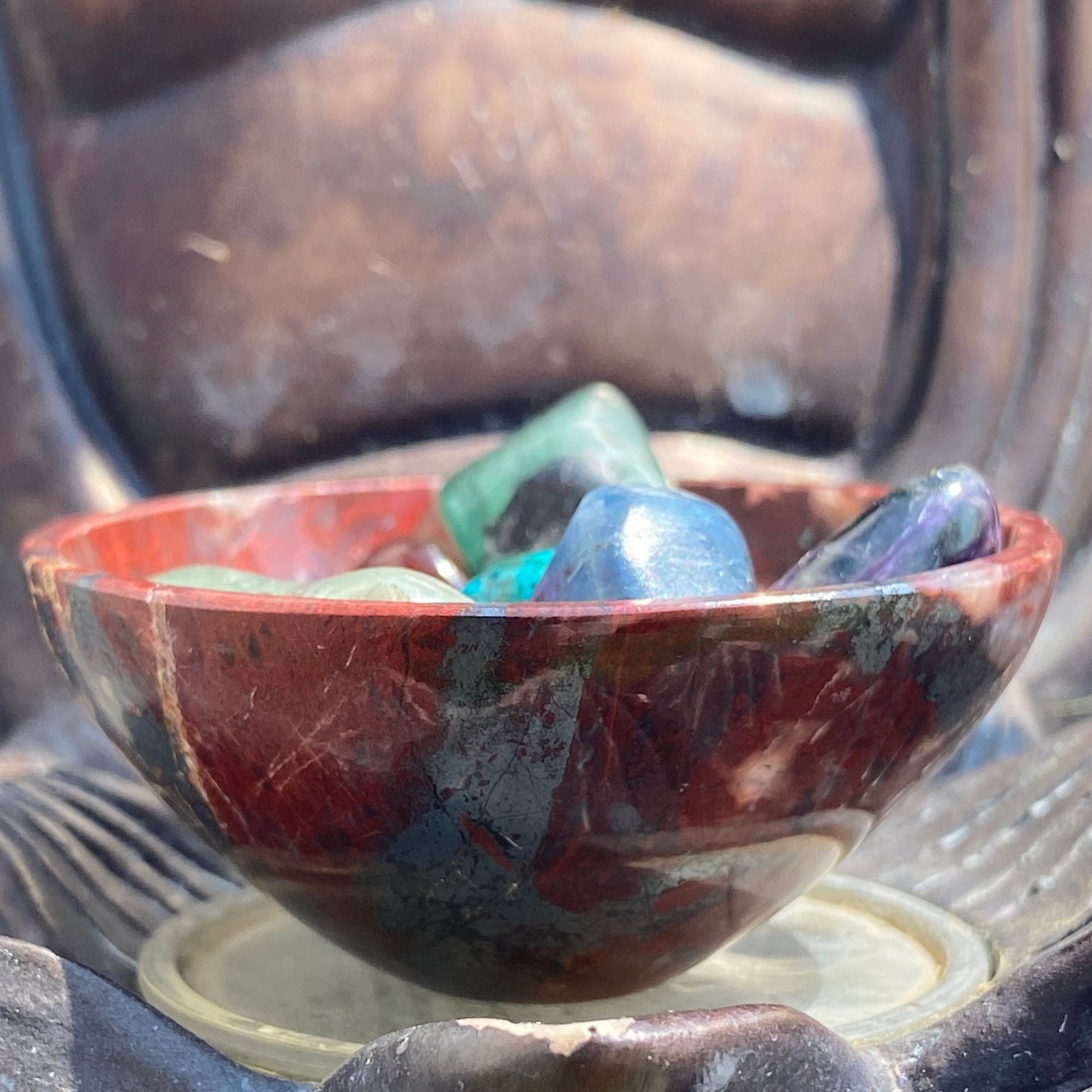 A close-up image of a polished 2" Mini Red Jasper Offering Bowl containing various tumbled gemstones in different colors, including green, blue, and purple. The bowl rests on a carved wooden surface with intricate details, possibly part of a larger sculpture or decorative piece used in spiritual practices.