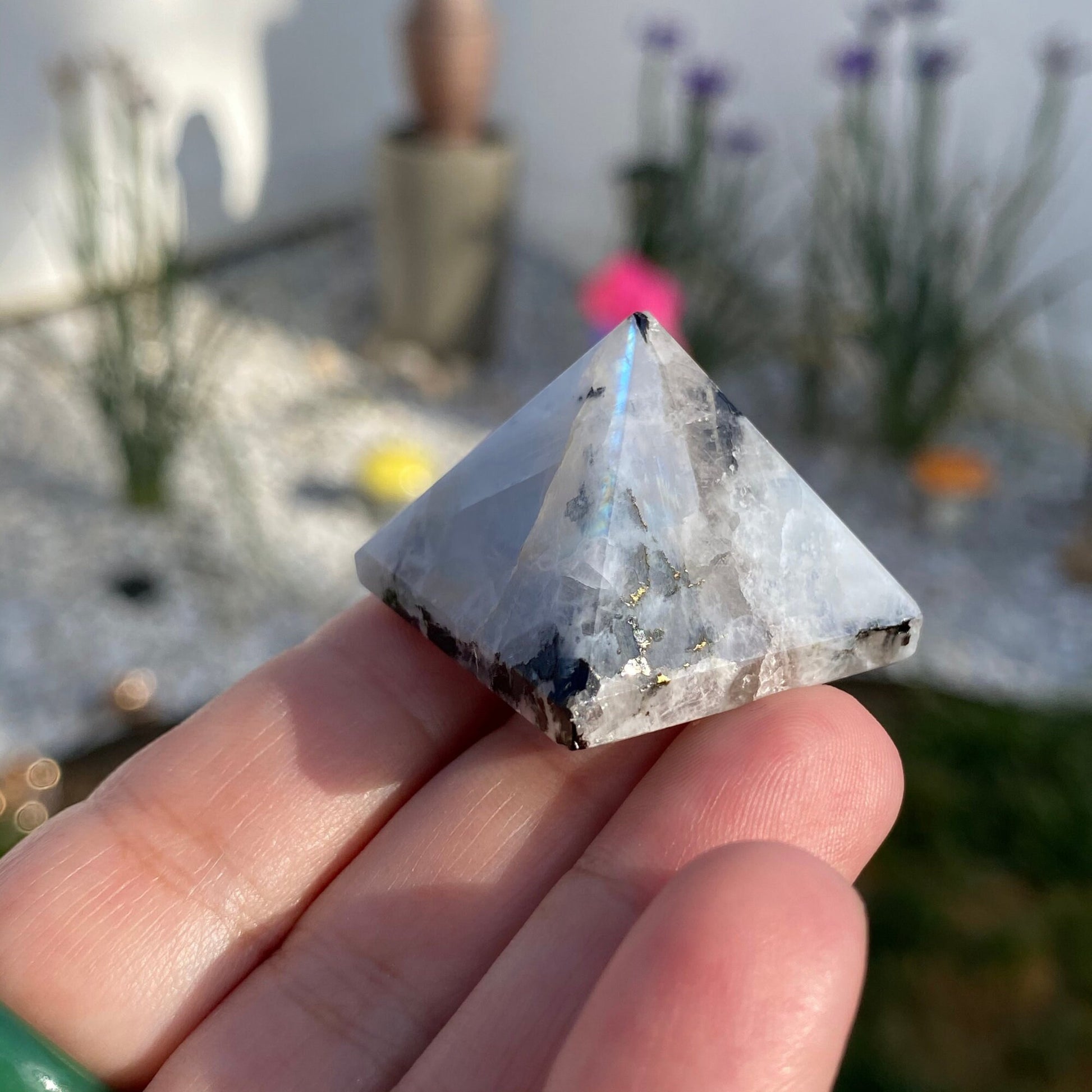 A close-up image of a person's hand with teal-painted nails, holding a 1" Mini Rainbow Moonstone Pyramid. The translucent stone, possibly a Rainbow Moonstone, features white and dark inclusions that reflect light brilliantly. The blurred outdoor background adds an ethereal touch to this scene.