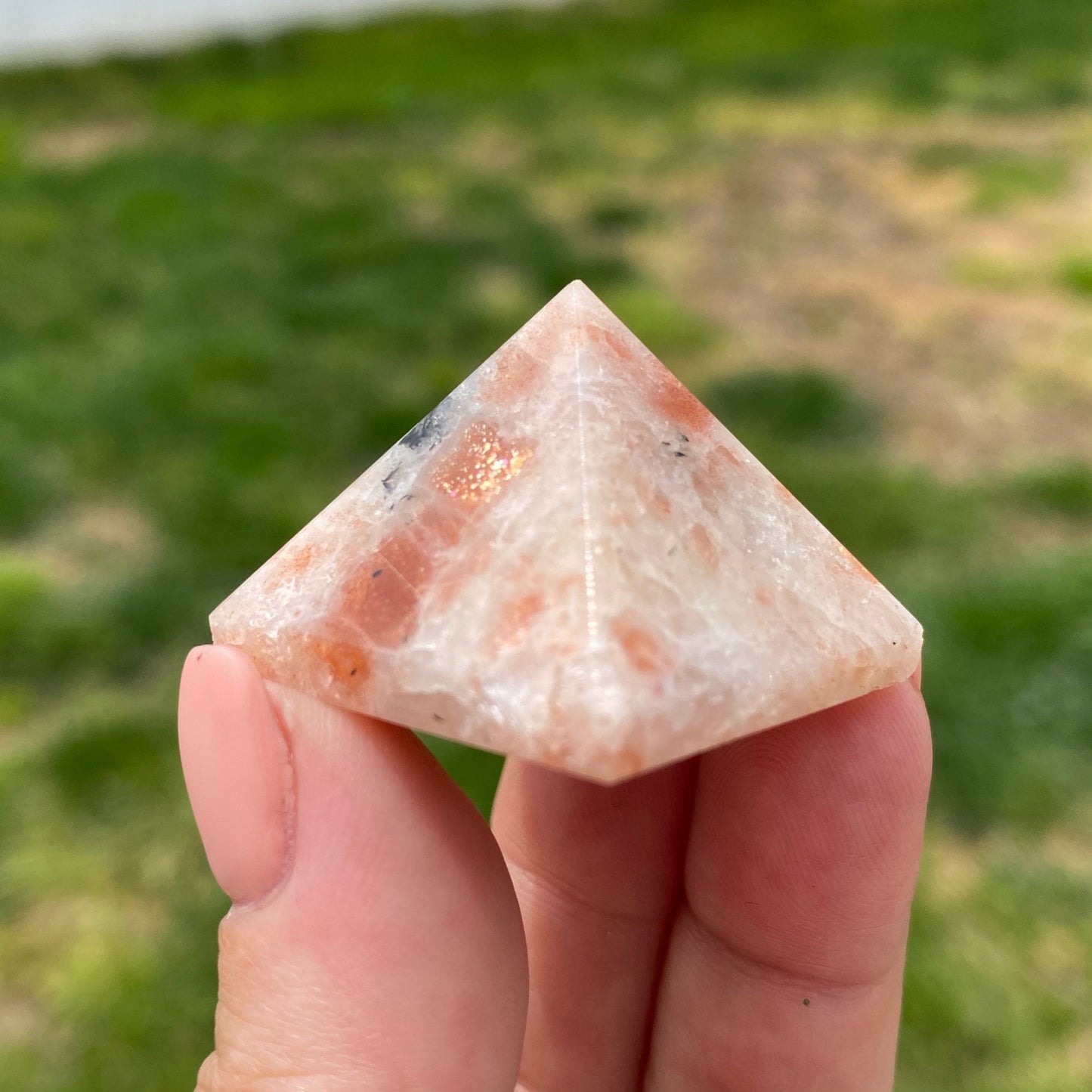 A hand with a manicured nail holds a small, polished 1" Mini Sunstone Pyramid with a mix of pink, white, and black colors. Known for bringing good fortune and self-empowerment, it is displayed against a grassy area with patches of dirt in the background.