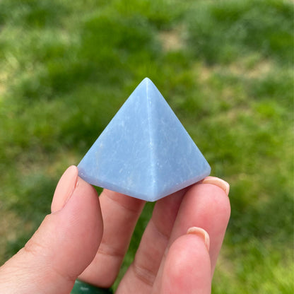 A hand holding a smooth, light blue 1" Mini Angelite Pyramid against a blurred green background. The pyramid has a shiny surface and distinct triangular faces, showcasing its polished and symmetrical structure, ideal for psychic healing.