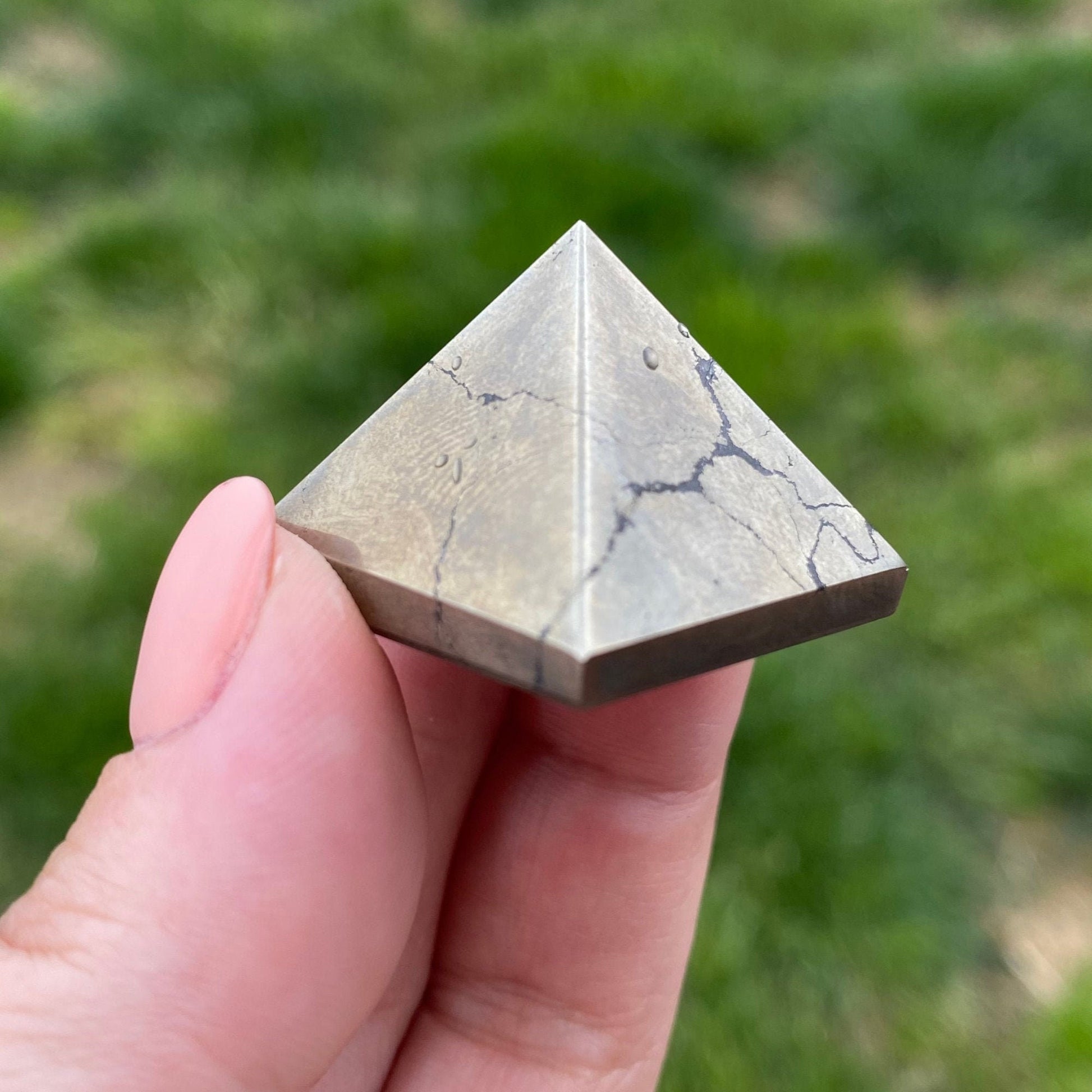 Three 1" Mini Pyrite Pyramids made of a dark stone are placed on a white surface in front of a white tiled background. Behind the pyramids, a bundle of dried sage and a piece of palo santo wood, known for their protective qualities, are set on the surface, aligned horizontally.