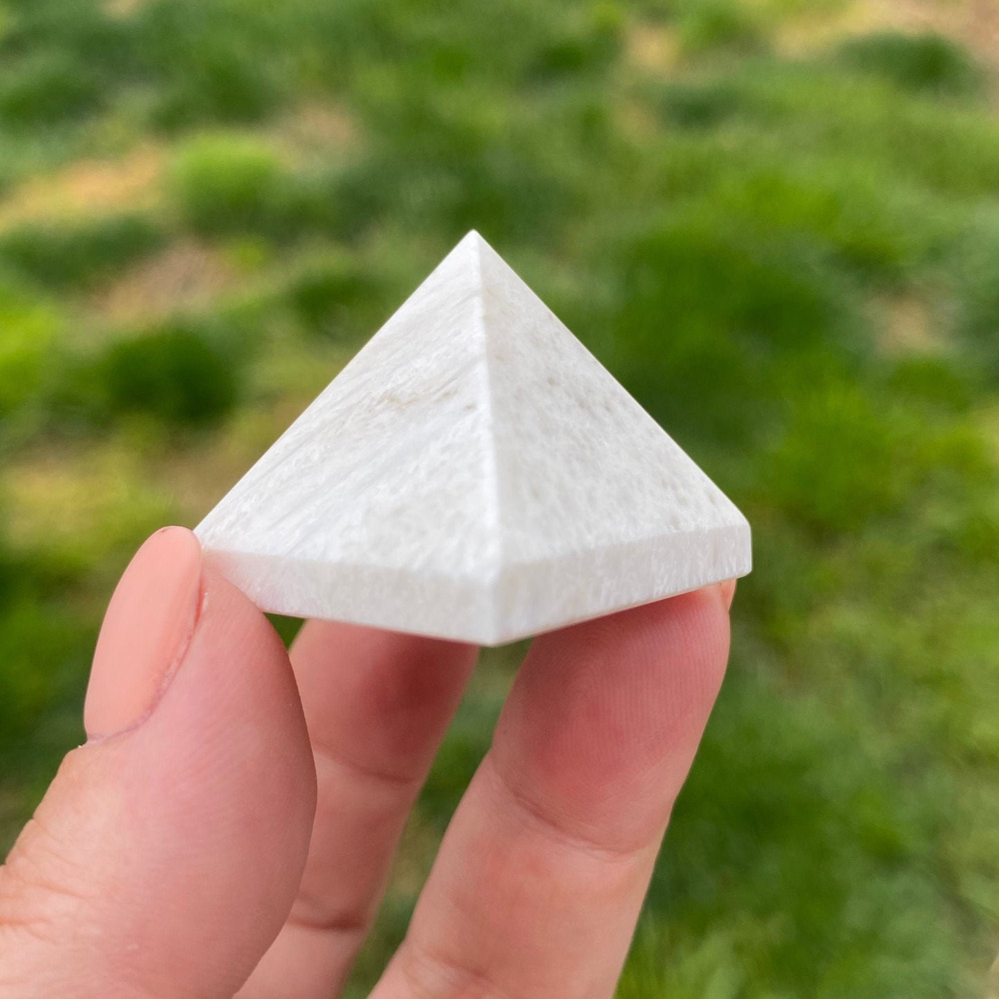 A hand holding a small, white 1" Mini Scolecite Pyramid outdoors with grass in the background, perfect as a meditation tool for achieving inner peace.