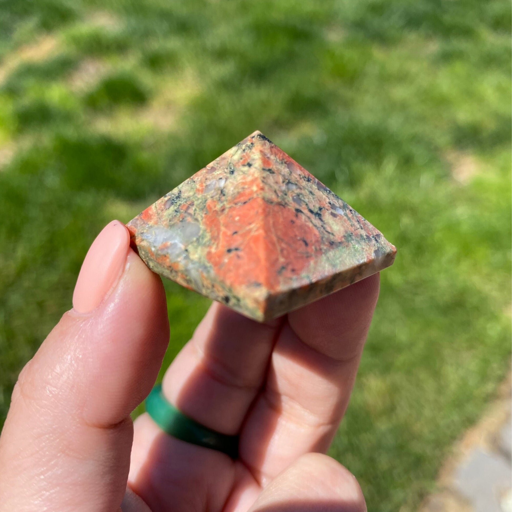 Close-up of a hand holding a 1" Mini Unakite Pyramid with a reddish-orange, green, and gray marbled pattern. The background is blurred greenery, emphasizing the stone's beauty and its potential for emotional balance.