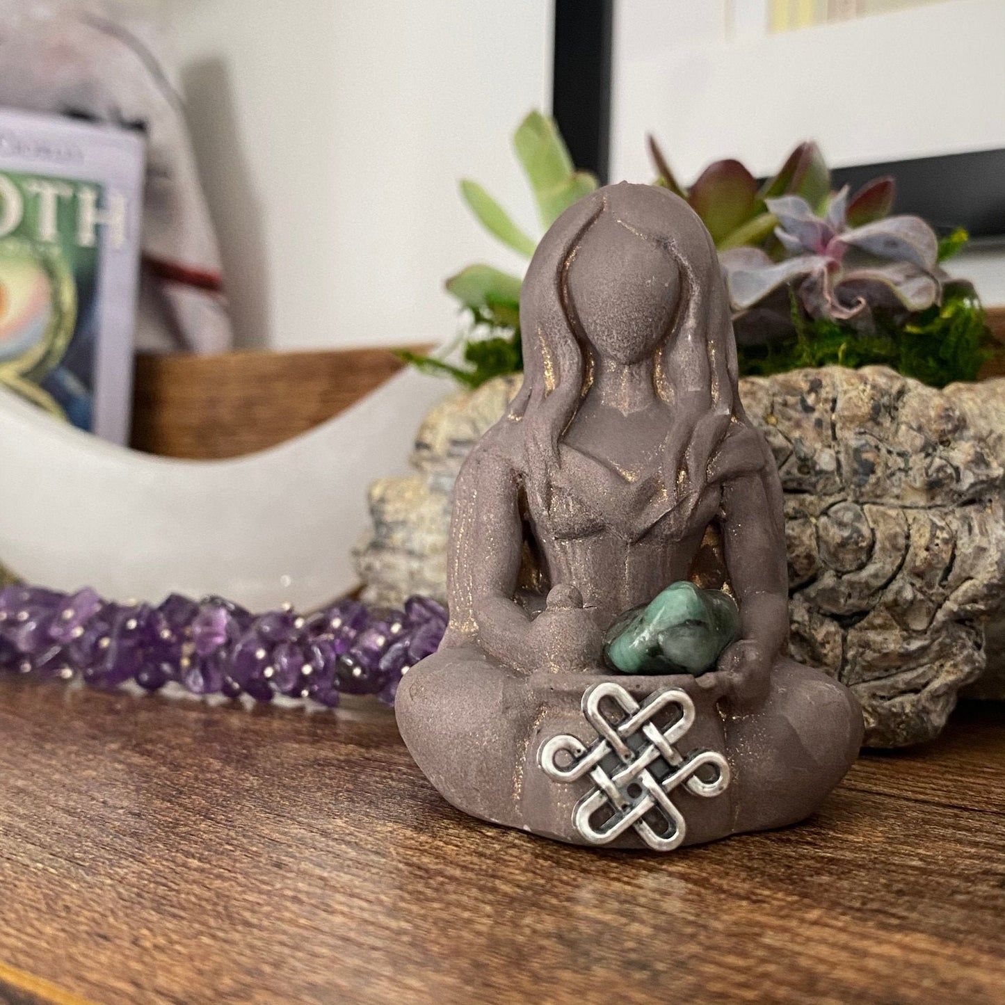 A stone Cerridwen Goddess Statue with long hair and no facial features sits cross-legged, holding an emerald pocket stone and a decorative knot. Behind it, a crescent moon ornament, a purple crystal bracelet, and various succulents are on display, radiating mystical energy.