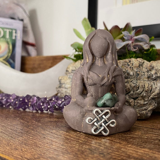 A stone Cerridwen Goddess Statue with long hair and no facial features sits cross-legged, holding an emerald pocket stone and a decorative knot. Behind it, a crescent moon ornament, a purple crystal bracelet, and various succulents are on display, radiating mystical energy.