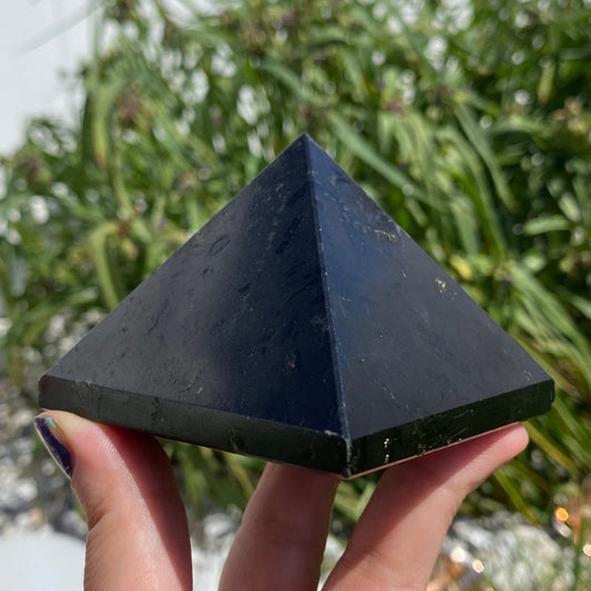 A hand holds a Large Black Tourmaline Crystal Pyramid, made of dark stone or crystal, with a green leafy plant visible in the blurred background—perfect for meditation and harnessing protective energy.