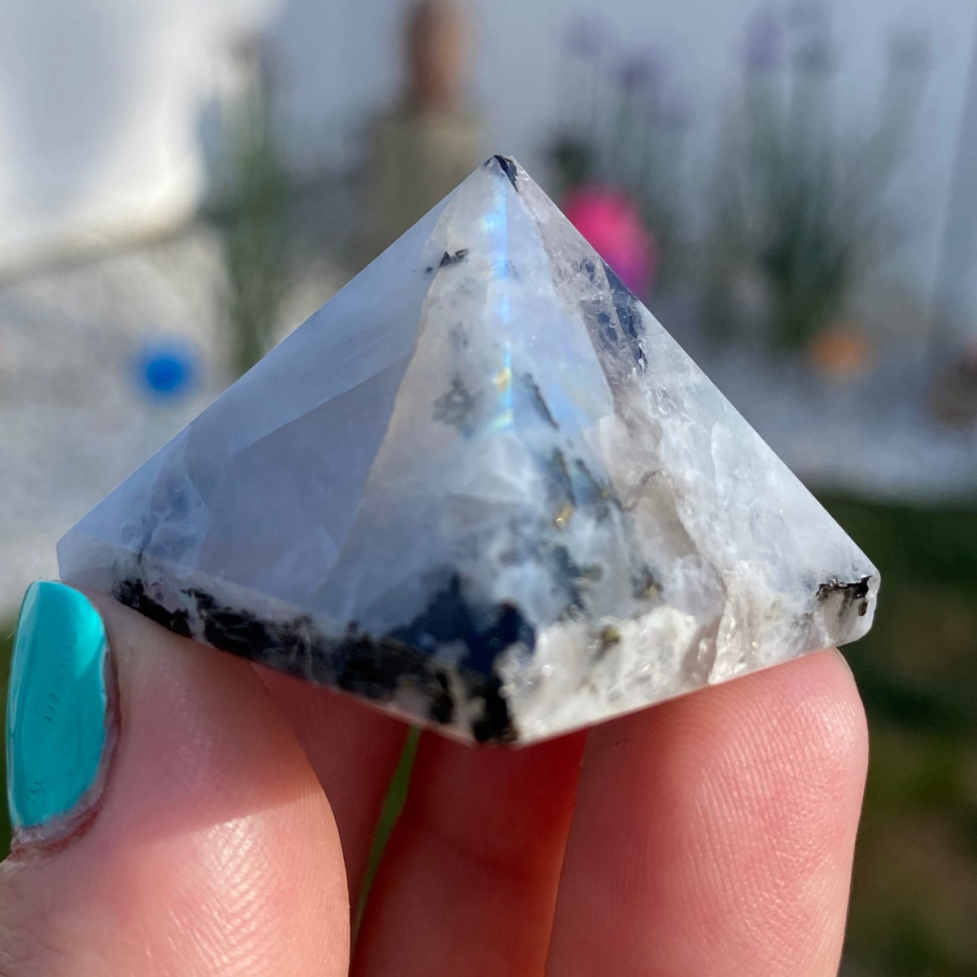 A close-up image of a person's hand with teal-painted nails, holding a 1" Mini Rainbow Moonstone Pyramid. The translucent stone, possibly a Rainbow Moonstone, features white and dark inclusions that reflect light brilliantly. The blurred outdoor background adds an ethereal touch to this scene.