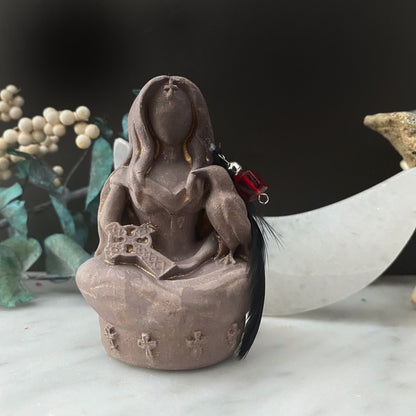 A sculpted figure with long hair, seated in a cross-legged meditation pose. The figure holds a cross in its lap, adorned with dark feathers reminiscent of a Morrigan Goddess Statue and a small red vial. Decorative elements, including white berries and a crescent moon, form the background.