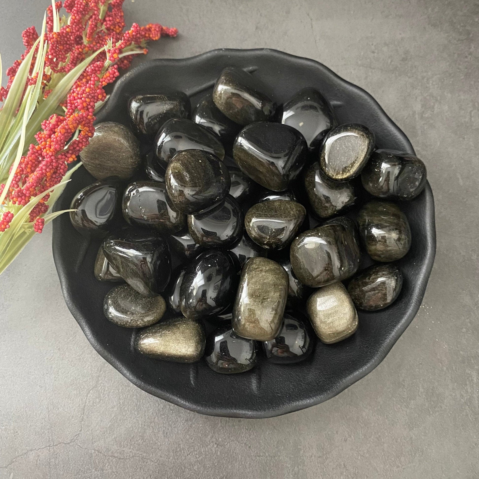 A black bowl filled with polished Gold Sheen Obsidian Tumbled Stones with metallic streaks. The bowl is placed on a grey surface with a bunch of red berries on stems lying beside it. The stones, known for psychic protection, have a smooth, shiny finish that reflects light beautifully.