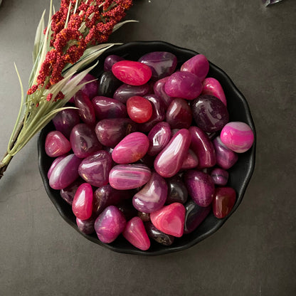 A black bowl filled with polished, colorful Pink Agate Tumbled Stones in various shades of pink and purple rests on a dark surface. Next to the bowl is a sprig of greenery with small red berries, highlighting the stones' healing properties and offering a touch of emotional support.