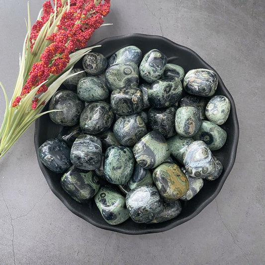 A black bowl filled with polished Kambaba Jasper Tumbled Stones, known as a calming stone, displays shades of green and black with swirling patterns. A sprig of green foliage and red berries lies beside the bowl on a gray surface.