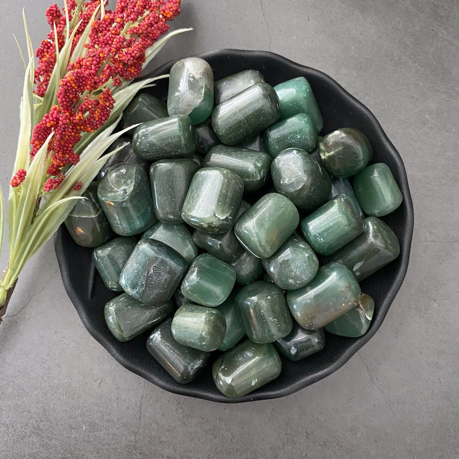 A black bowl filled with polished, rounded Bowenite Tumbled Stones sits on a dark gray surface. To the left of the bowl is a sprig of red berries and green foliage, adding a touch of contrast to the image and highlighting the protective qualities believed to aid in dream recall.