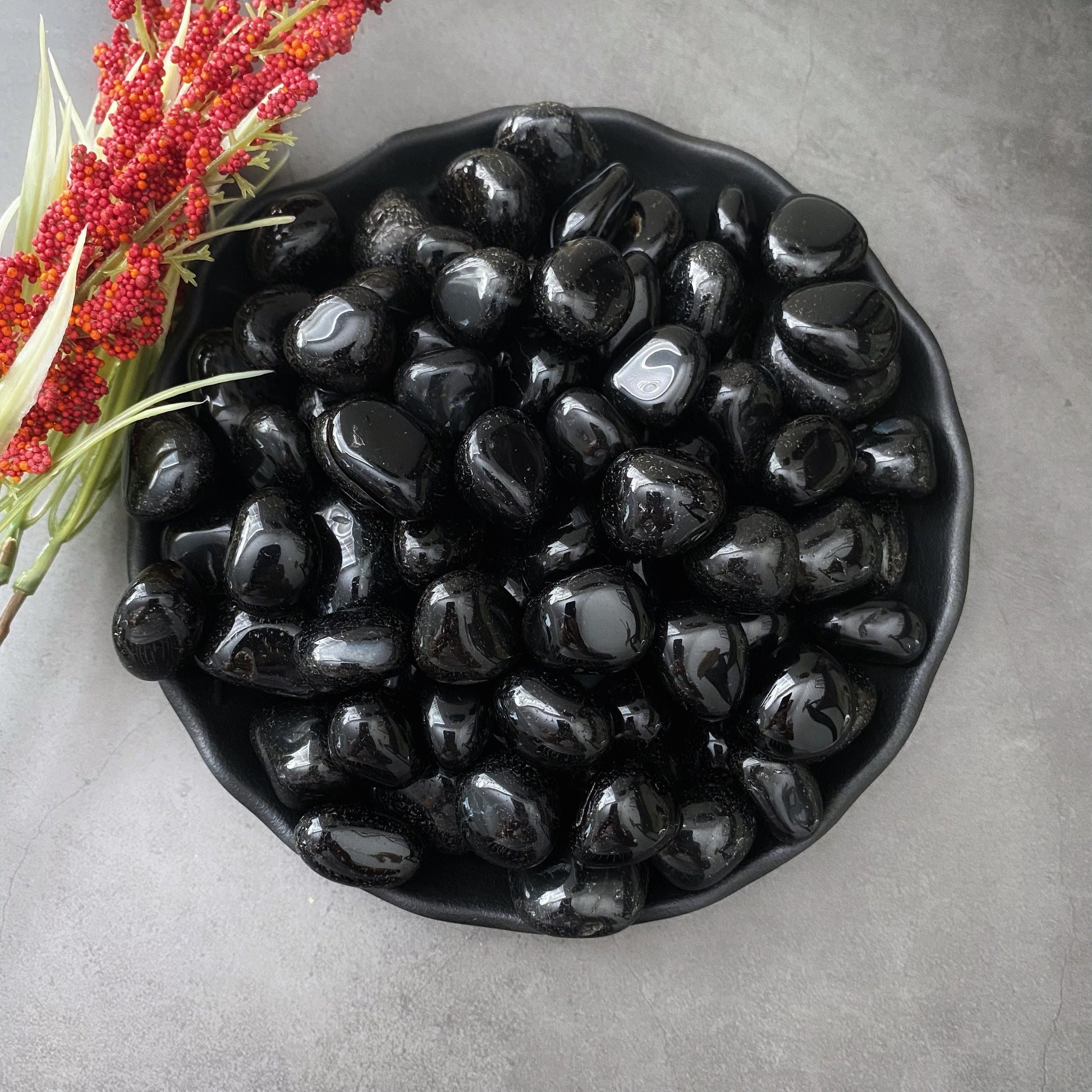 A black ceramic bowl filled with smooth, shiny Black Onyx Tumbled Stones sits on a gray surface. To the left of the bowl are sprigs of red and white artificial flowers, adding contrast to the monochrome setting. This arrangement serves as a subtle protection amulet for enhancing spiritual vision.