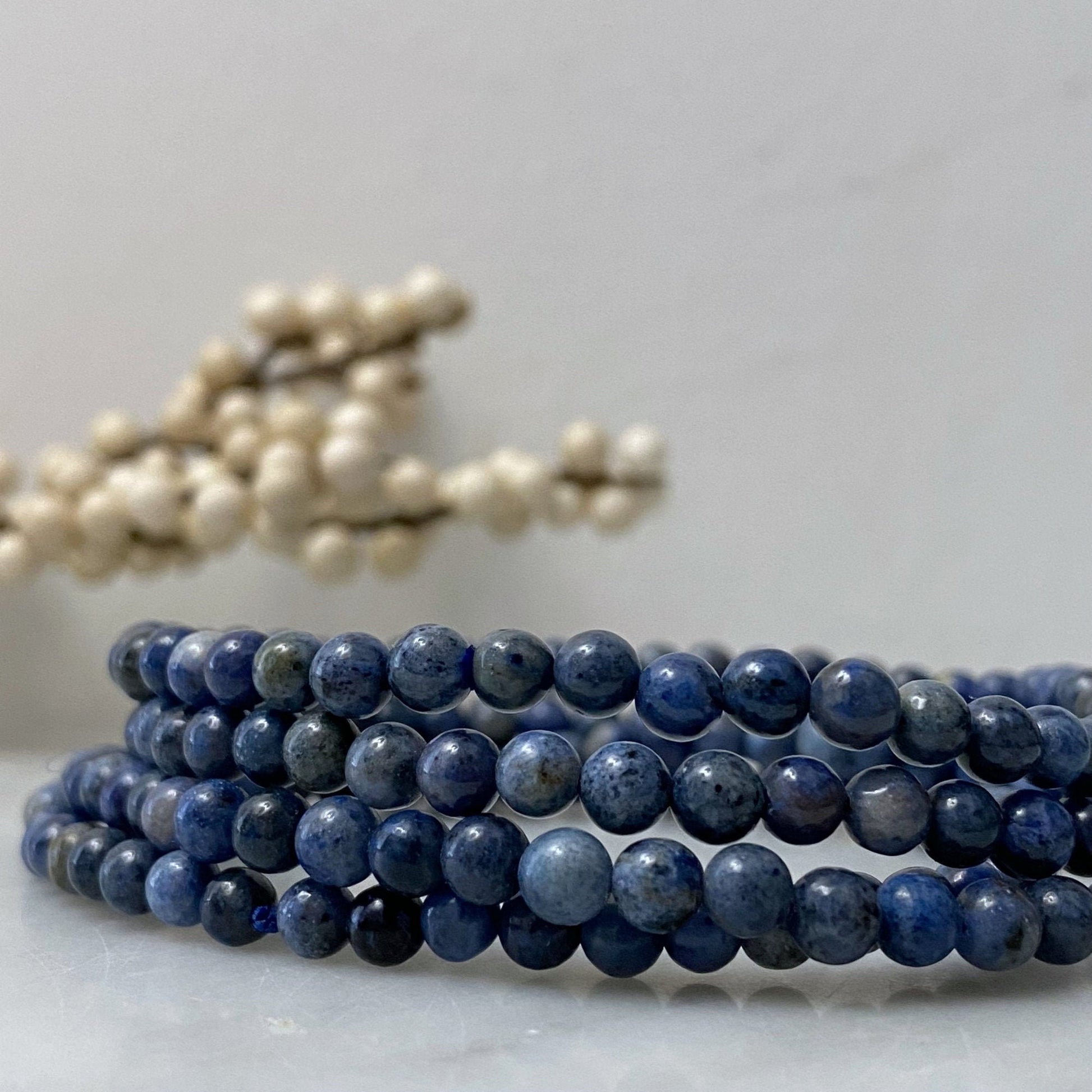 A close-up photo of multiple strands of round, polished 4mm Dumortierite Beaded Bracelets coiled together. In the background, a blurred bunch of white spherical objects resembling berries on stems is situated on a light-colored surface. The scene exudes calming Usui Reiki energy.