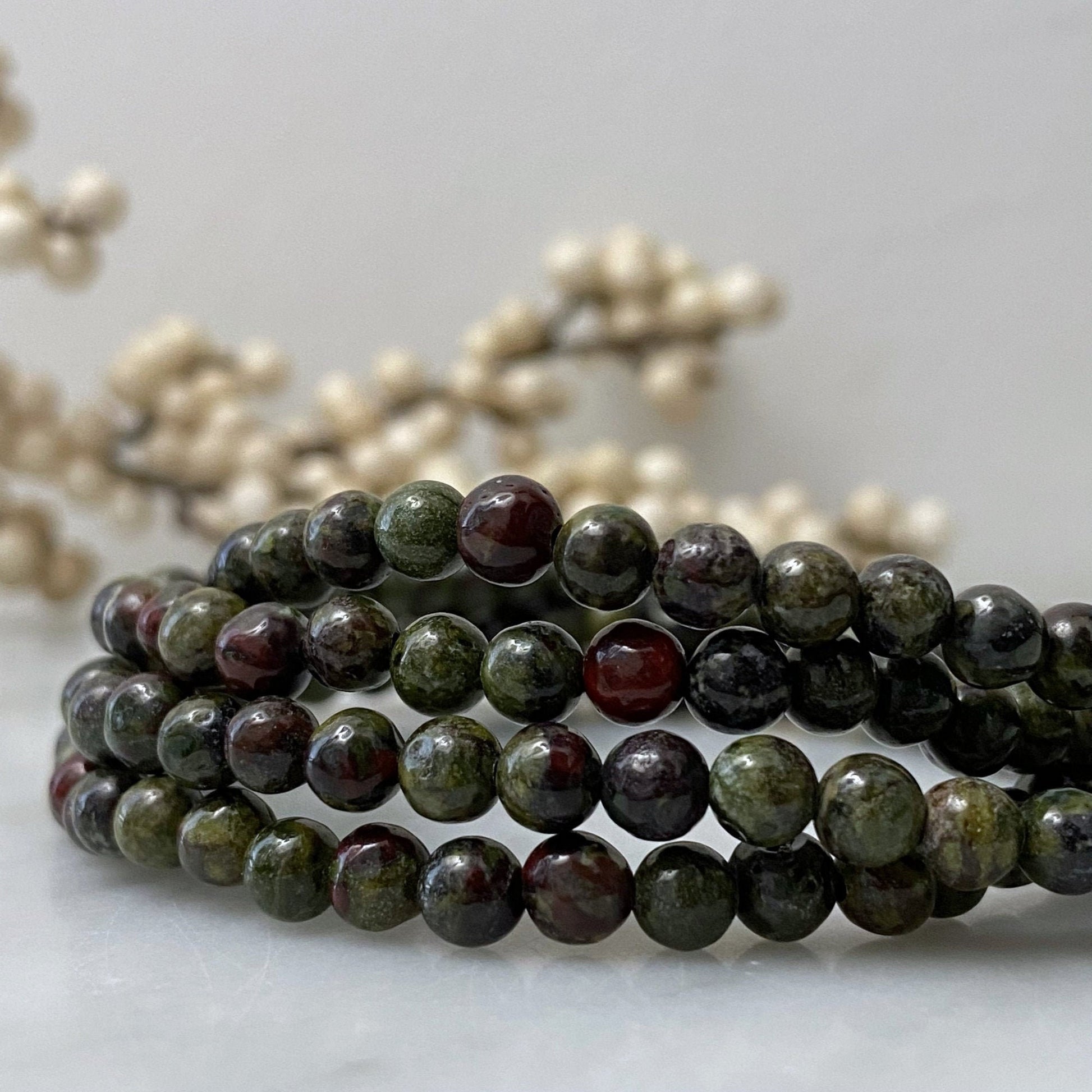 A close-up of a 4mm Dragon Stone Beaded Bracelet made of dark, round, polished beads with varying shades of brown and green. In the background, there is an out-of-focus arrangement of white berries, enhancing the bracelet's natural and earthy aesthetics.