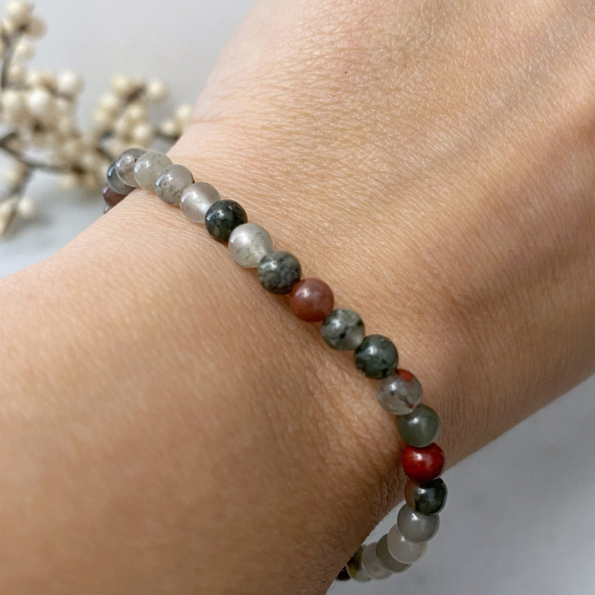 A close-up image of a stretchy, multi-strand 4mm Seftonite (African Bloodstone) Beaded Bracelet made of semi-transparent stones in various colors, including green, red, and grey. The bracelet is coiled neatly, exuding vibrant Reiki energy against a blurred backdrop featuring white, pearl-like beads.