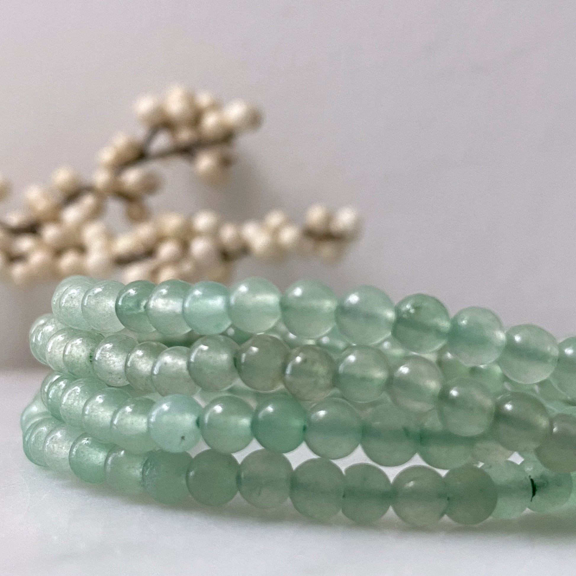 A delicate 4mm Green Aventurine Beaded Bracelet made of polished green beads is centered against a black background. The uniform beads highlight its stretchable design, and the bracelet's reflection shimmers below it on the reflective surface.