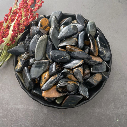 A black bowl filled with polished Blue Tiger Eye Tumbled Stones, featuring slight brown and gray streaks. The bowl is set on a dark gray surface, with red dried flowers displayed next to it, adding a touch of color and enhancing the soothing stone composition for stress and anxiety reduction.