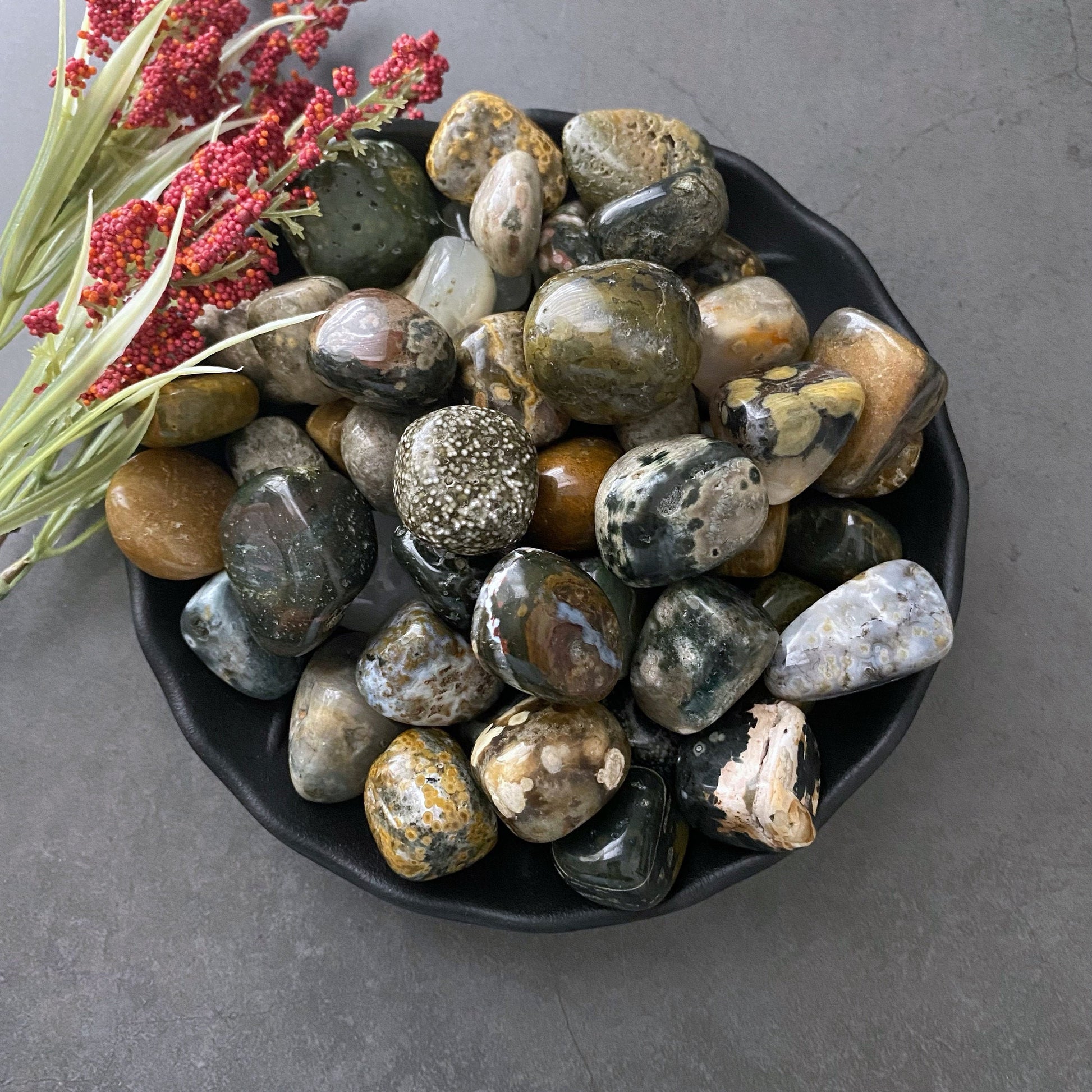 A black bowl filled with a variety of polished, multicolored stones and pebbles, including Ocean Jasper Tumbled Stones for emotional stability, sits on a gray surface. To the left of the bowl, there is a small arrangement of red flowers and green foliage.