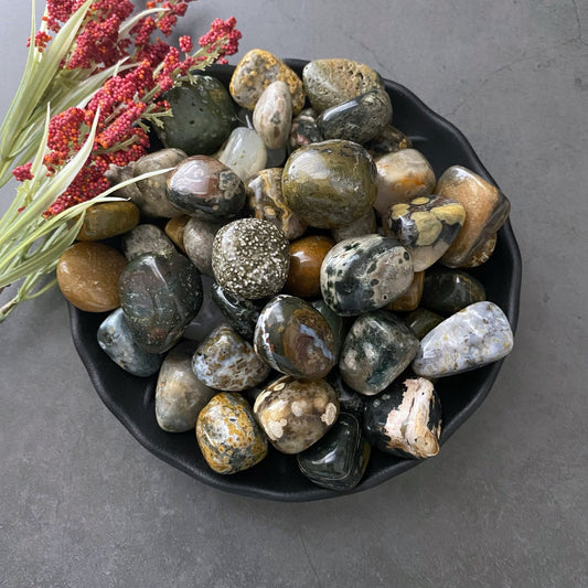 A black bowl filled with a variety of polished, multicolored stones and pebbles, including Ocean Jasper Tumbled Stones for emotional stability, sits on a gray surface. To the left of the bowl, there is a small arrangement of red flowers and green foliage.