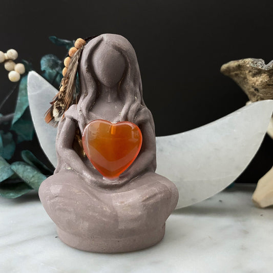 A serene, faceless Aine Goddess Statue sits cross-legged, holding an orange heart-shaped gem. The Celtic Goddess statue features feather details on its side and is surrounded by greenery and small decorative elements, including a large crescent moon shape in the background.