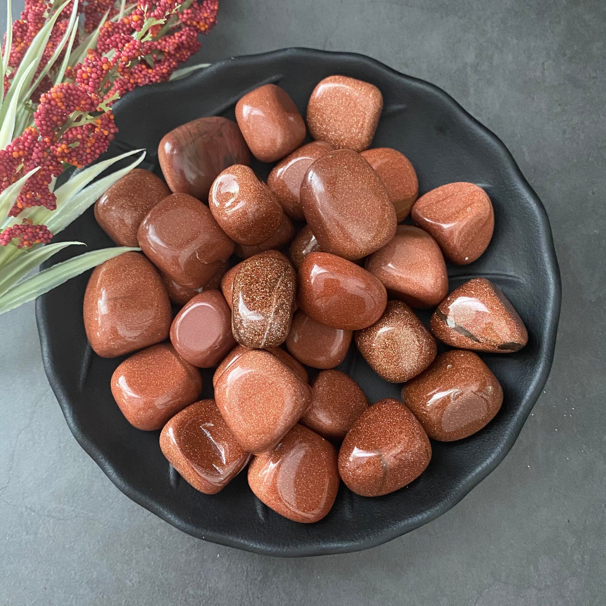 A black ceramic plate holding several polished, reddish-brown Goldstone Tumbled Stones is placed on a dark gray surface. Beside the plate is a small bouquet of red and green sprigs, enhancing the natural beauty and metaphysical properties of this healing stone composition.