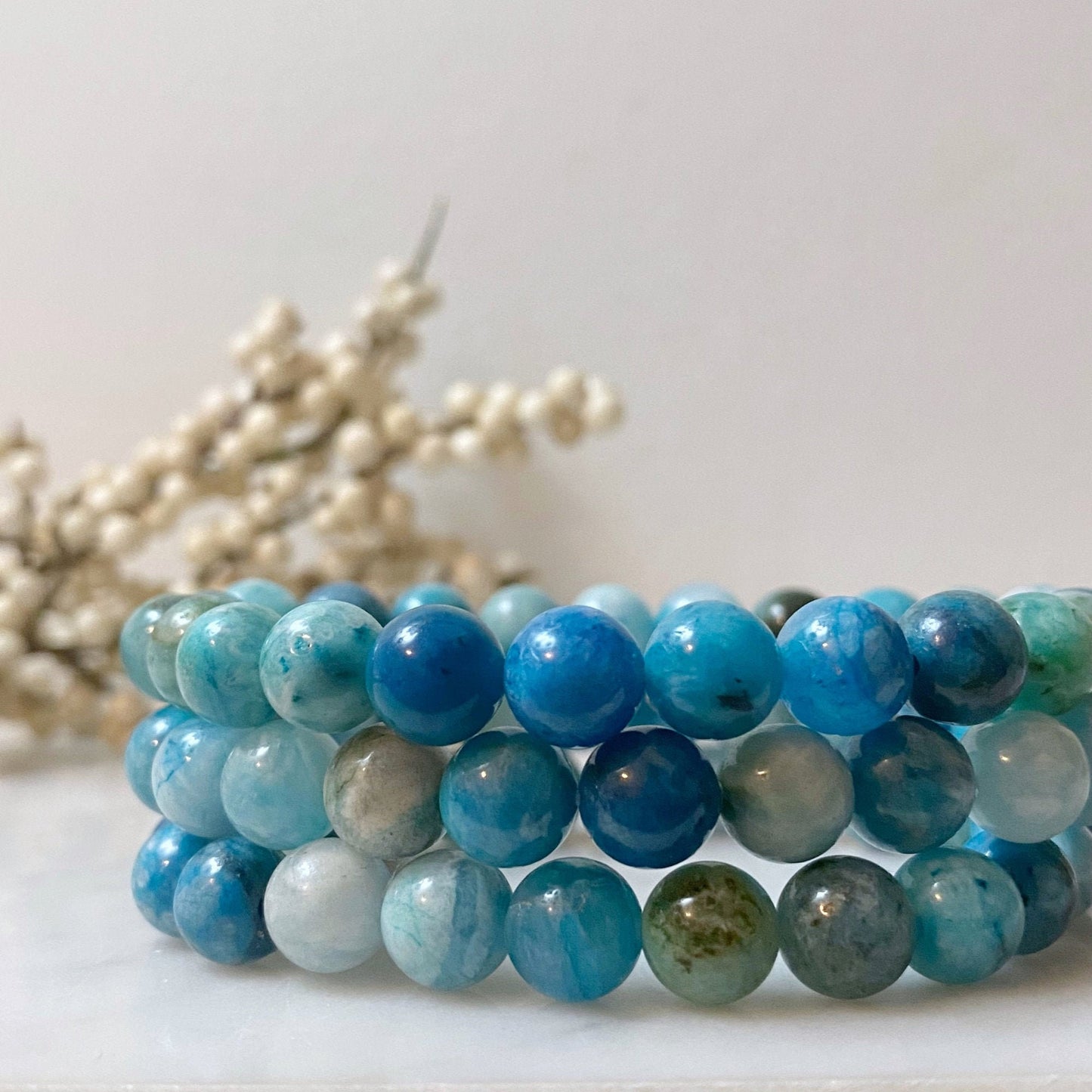 A stunning 8mm Hemimorphite Beaded Bracelet made of round, polished blue stones is displayed against a black background. The stones vary in shades of blue and exhibit slight marbling, emanating Reiki energy. The bracelet's reflection shimmers on the shiny surface beneath it.