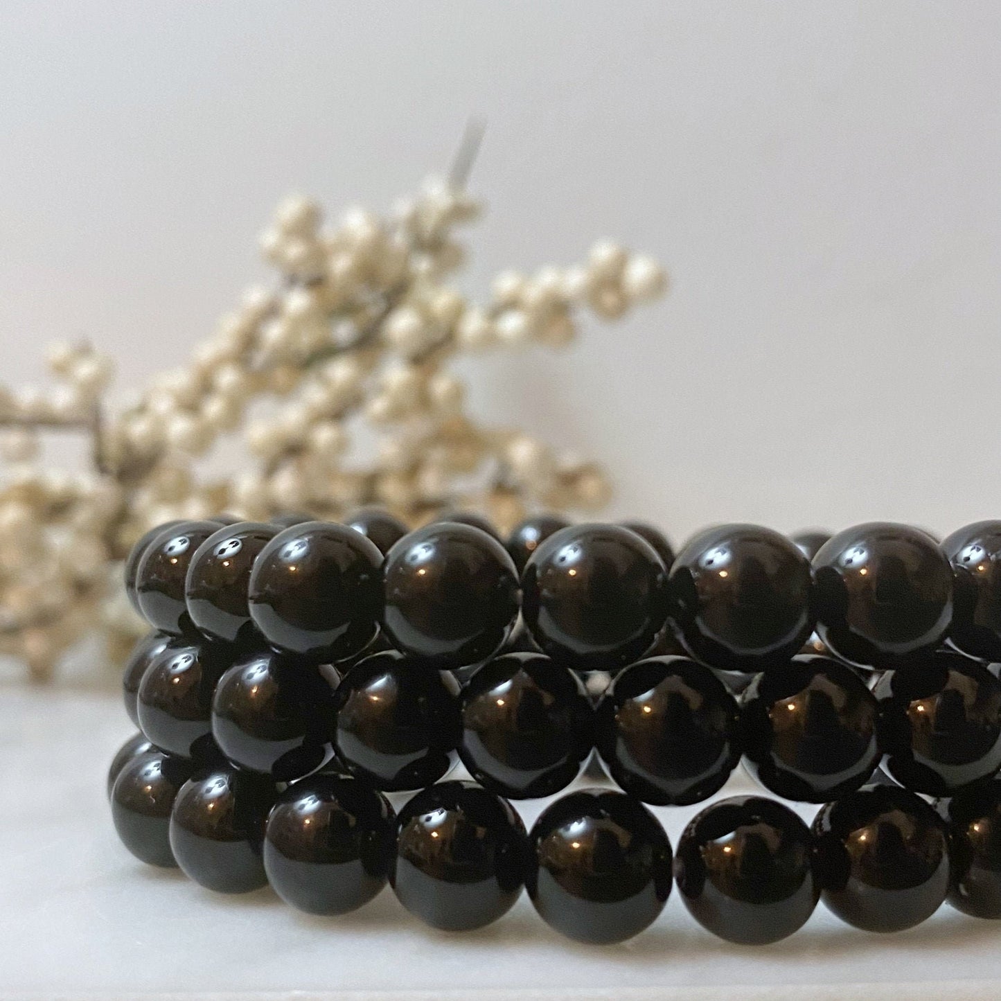 A beautiful 8mm Black Onyx Beaded Bracelet made of glossy black beads is displayed on a white surface. In the background, out of focus, there's a delicate branch with small white berries. The composition creates a pleasing contrast between the shiny beads and the soft white berries, emanating psychic protection.