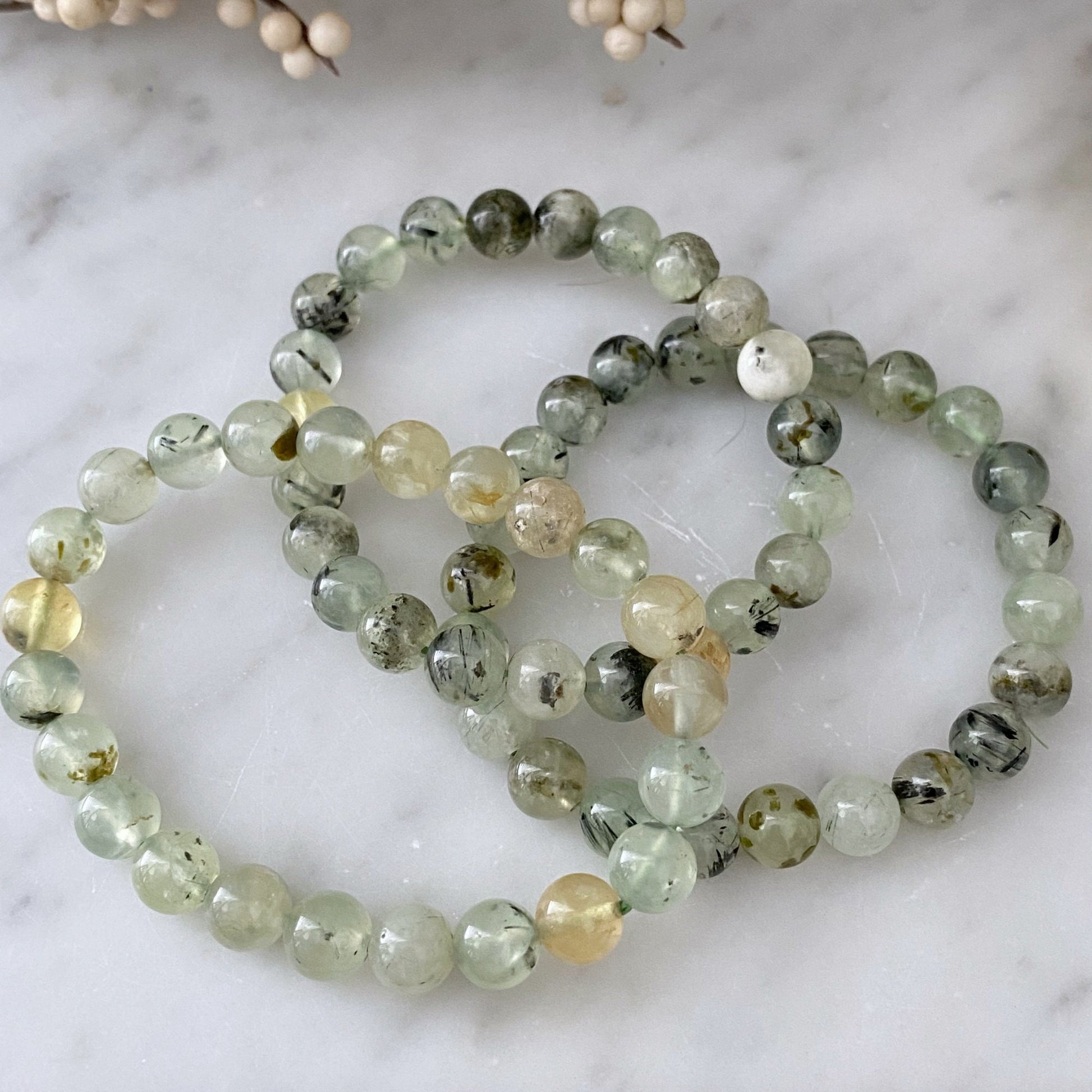 A close-up shot of an 8mm Prehnite Beaded Bracelet made from translucent green beads with dark inclusions. The bracelet is coiled in multiple loops on a light grey surface. In the background, a blurred branch with small, off-white buds is visible, creating a soft, natural ambiance.