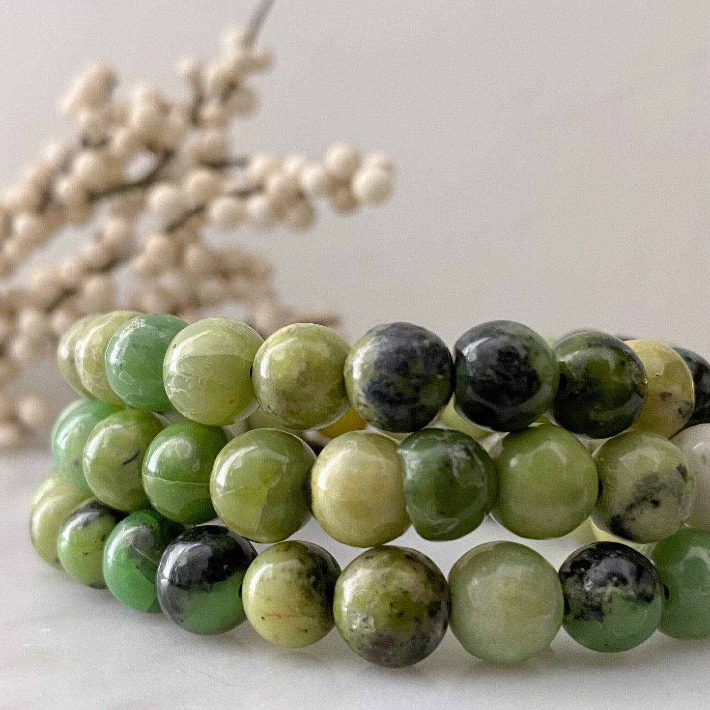 Close-up image of three beaded bracelets made from green and black semi-precious stones, including an 8mm Chrysoprase Beaded Bracelet. The bracelets are stacked together on a light-colored surface, with a blurred background featuring off-white berries on delicate branches.