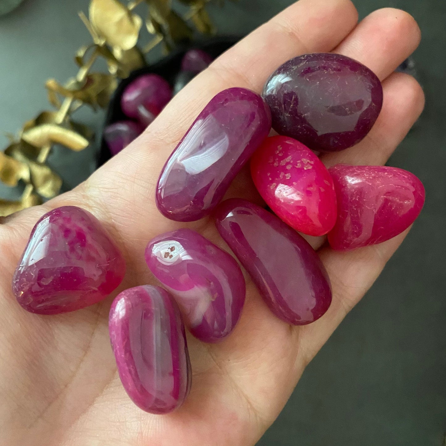 A black bowl filled with polished, colorful Pink Agate Tumbled Stones in various shades of pink and purple rests on a dark surface. Next to the bowl is a sprig of greenery with small red berries, highlighting the stones' healing properties and offering a touch of emotional support.