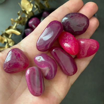 A black bowl filled with polished, colorful Pink Agate Tumbled Stones in various shades of pink and purple rests on a dark surface. Next to the bowl is a sprig of greenery with small red berries, highlighting the stones' healing properties and offering a touch of emotional support.