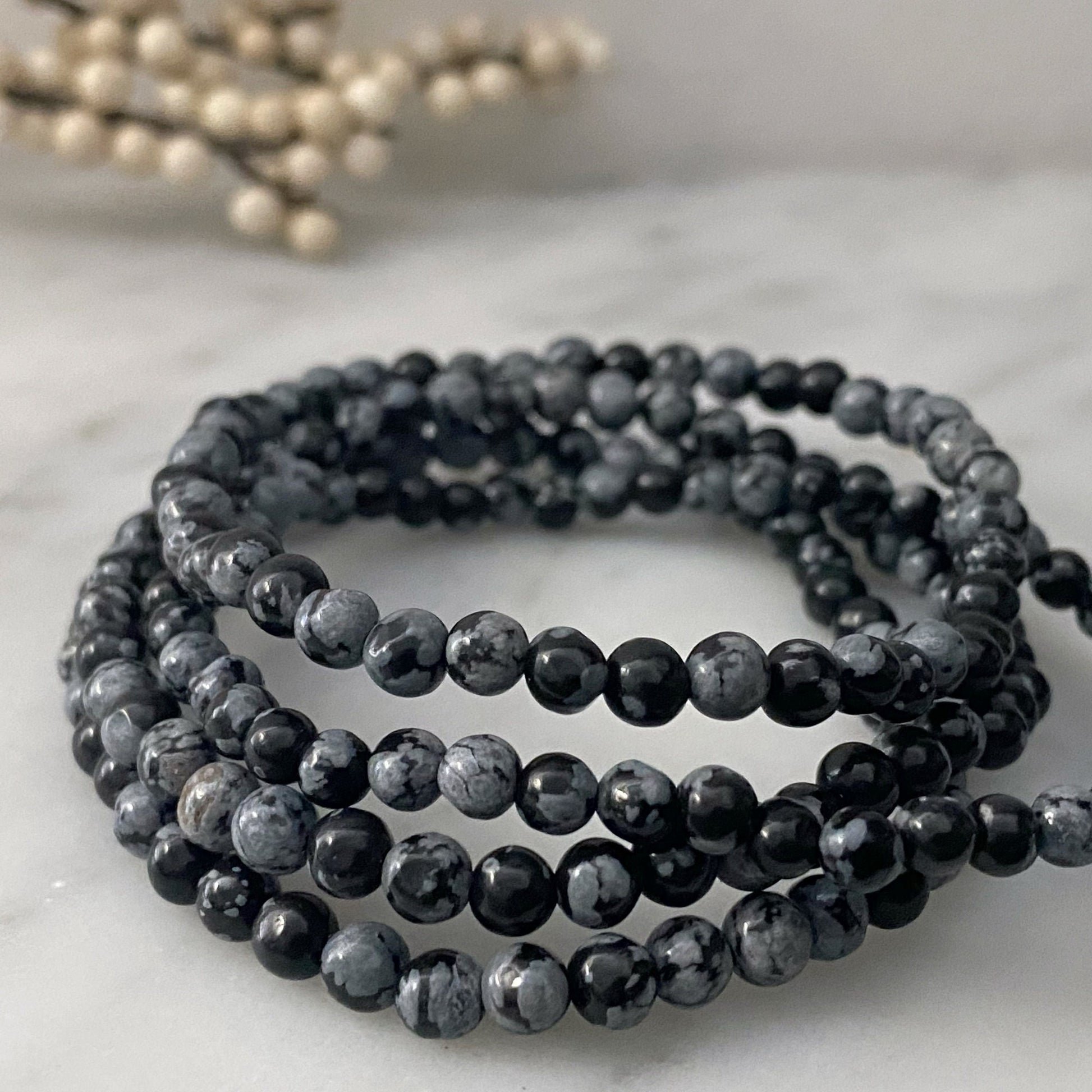 An image shows several strands of black and gray marbled 4mm Snowflake Obsidian Beaded Bracelets coiled together on a white surface. In the background, there are out-of-focus cream-colored berries on a branch, adding a decorative element to the composition.