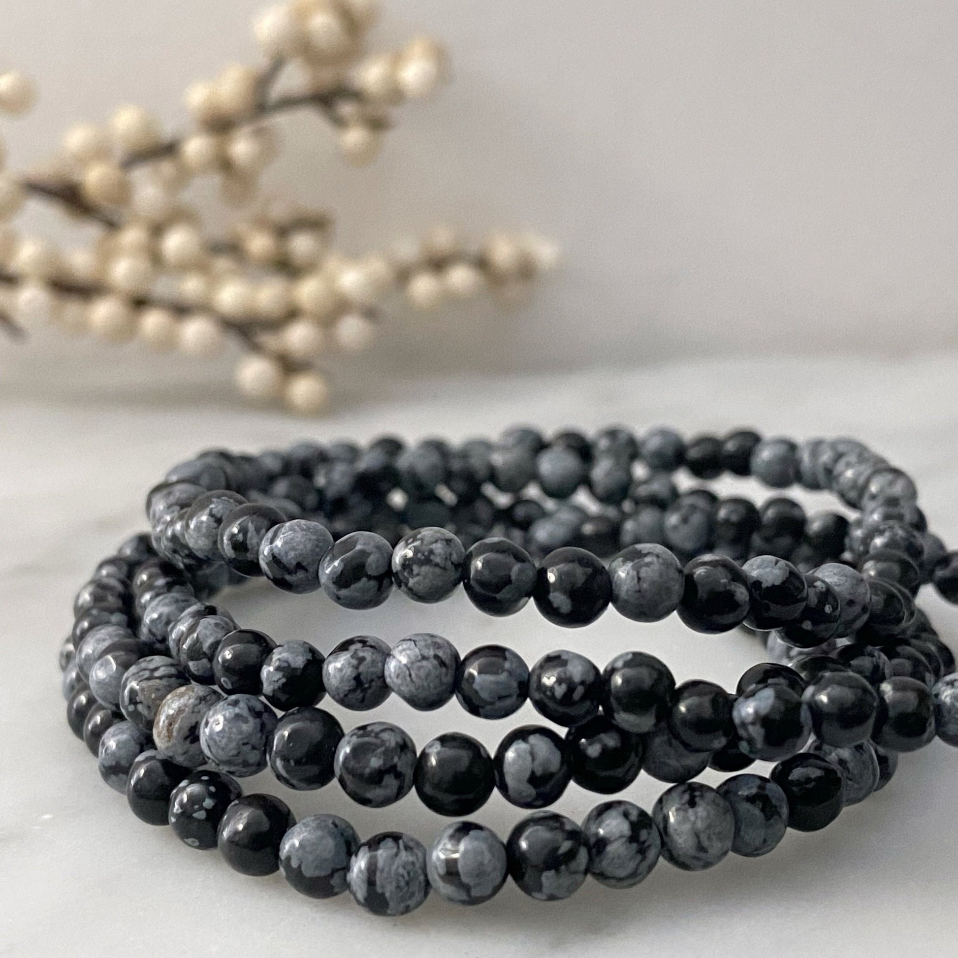 An image shows several strands of black and gray marbled 4mm Snowflake Obsidian Beaded Bracelets coiled together on a white surface. In the background, there are out-of-focus cream-colored berries on a branch, adding a decorative element to the composition.