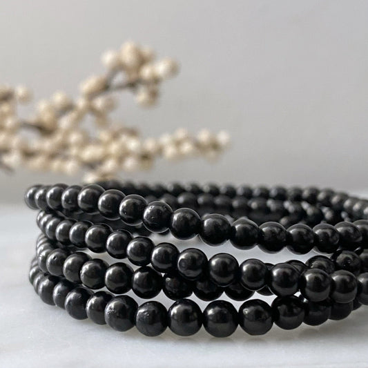 A stack of elegant black 4mm Jet Beaded Bracelets is arranged in a neat circle on a light marble surface. The background features soft-focus white berries, enhancing the simple yet sophisticated aesthetic of the scene while offering the grounding and protection stone's natural allure.