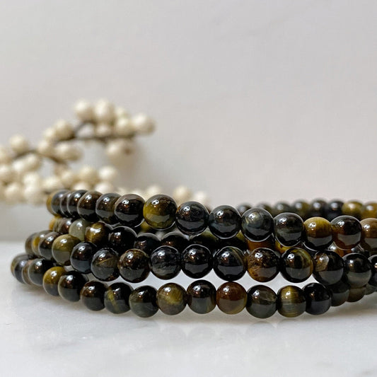 A close-up image of a 4mm Blue Tiger's Eye Beaded Bracelet, made of polished brown and black beads intricately wrapped around in multiple loops. The background is soft and out of focus, with light-colored decorative branches subtly visible, exuding a soothing stone energy.