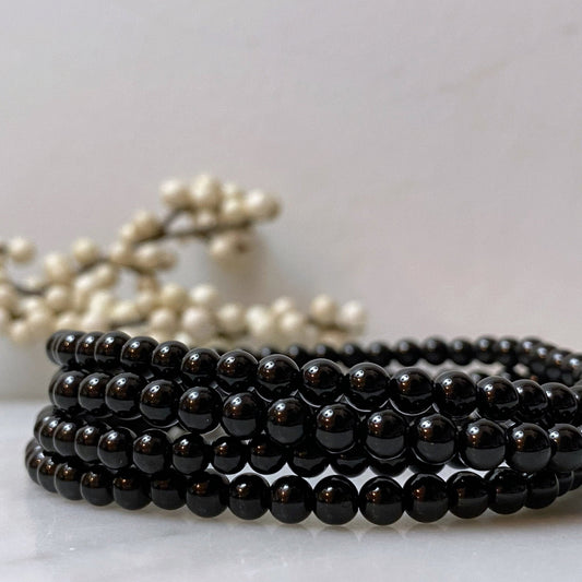 A set of four 4mm Black Tourmaline Beaded Bracelets is neatly stacked together in front of a delicate white and brown branch decoration. The glossy surface of the beads reflects light, adding a subtle shine while highlighting their purifying properties against the otherwise matte background.