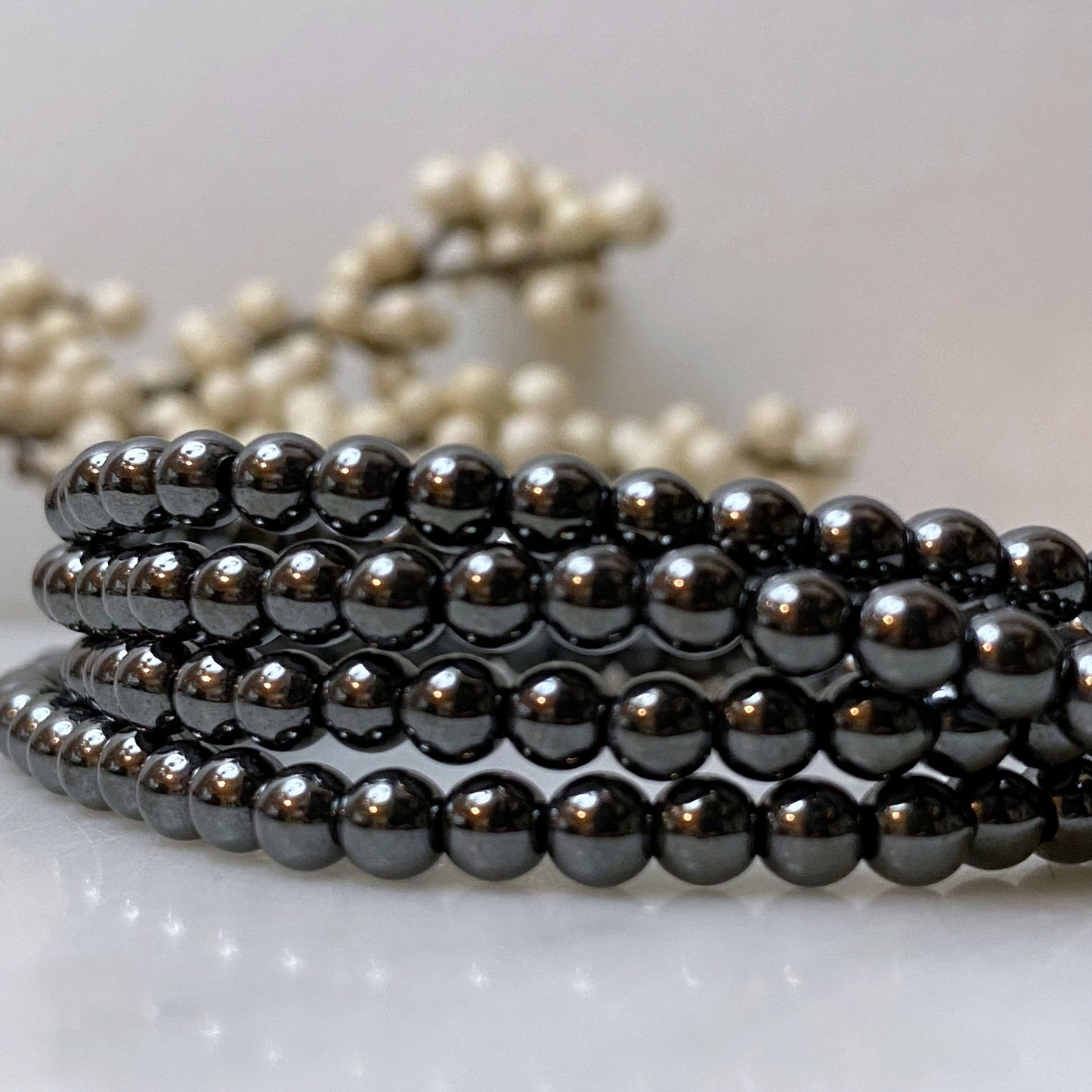 A close-up of a 4mm Hematite Beaded Bracelet made of shiny, reflective black beads. In the blurred background, there are small white spherical decorative elements. The overall tone of the image is elegant and minimalist, highlighting the grounding properties of the polished hematite beads.